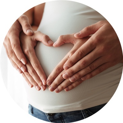 Mãos de um casal em forma de coração sobre barriga grávida com camisola branca e jeans azuis