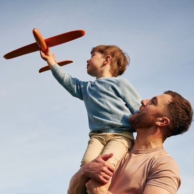 Menino segurando avião de brinquedo laranja no céu, apoiado nos ombros do homem.