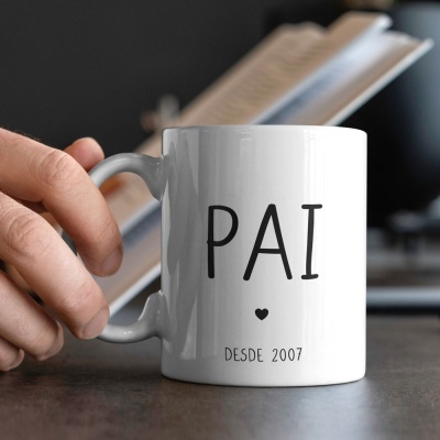 Caneca branca com texto 'PAI' e 'DESDE 2007', mão segurando-a, livro ao fundo