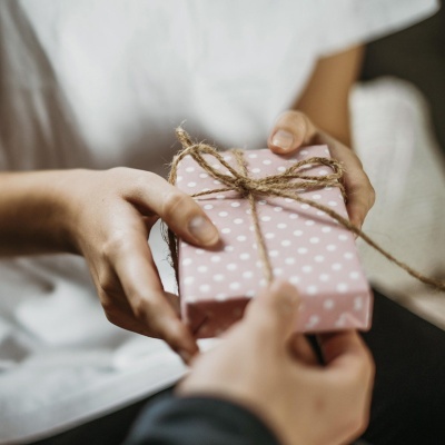 Pacote de presente rosa com bolinhas brancas e cordão de juta sendo entregue por duas mãos
