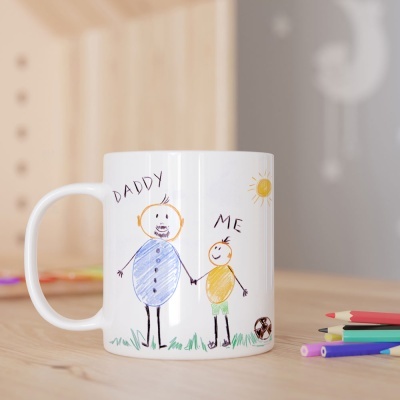 Caneca branca com desenho infantil de pai e filho em cores, sobre mesa de madeira com lápis coloridos ao lado.