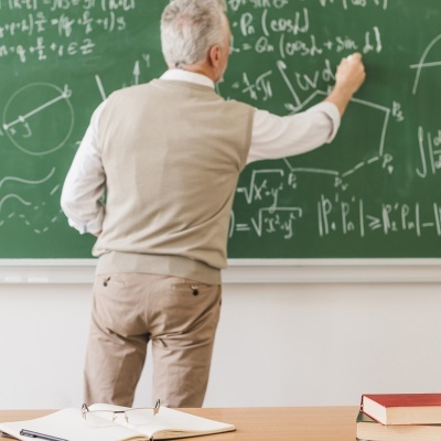 Professor escrevendo fórmulas num quadro verde com materiais de estudo numa mesa