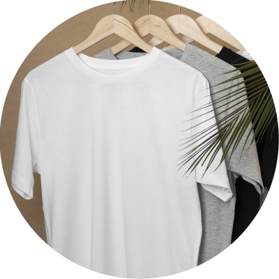Três t-shirts básicas branca, cinza e preta penduradas em cabides de madeira com planta ao lado