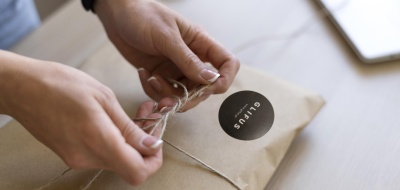 Pacote de papel castanho a ser amarrado com corda rústica por mãos femininas.