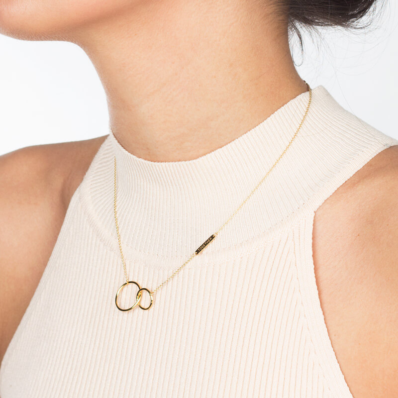 Omnia Colar Elos dourado. Colar dourado com dois círculos entrelaçados numa blusa creme de gola alta