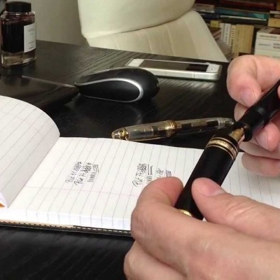 Pessoa segurando caneta preta com detalhes dourados junto a caderno aberto numa mesa preta com rato, teclado e frasco de tinta ao fundo