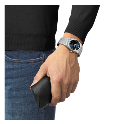 Relógio metálico com mostrador azul no pulso esquerdo e estojo preto na mão de pessoa com camisola preta e jeans azul