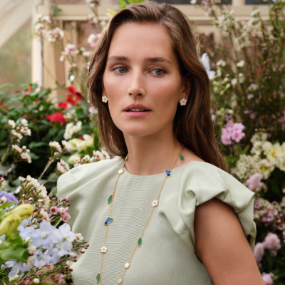 Mulher com brincos e colar florais, vestido verde, entre flores coloridas em estufa