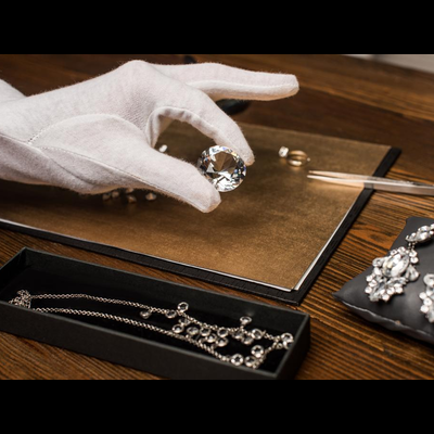 Mão com luva branca segura pedra preciosa transparente sobre bancada com joias prateadas e livro marrom.