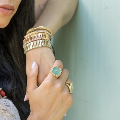 Anéis e pulseiras douradas com pedras verde e elementos salmão na mão e braço de uma mulher.