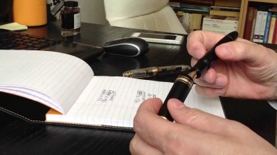 Pessoa segurando caneta preta com detalhes dourados junto a caderno aberto numa mesa preta com rato, teclado e frasco de tinta ao fundo