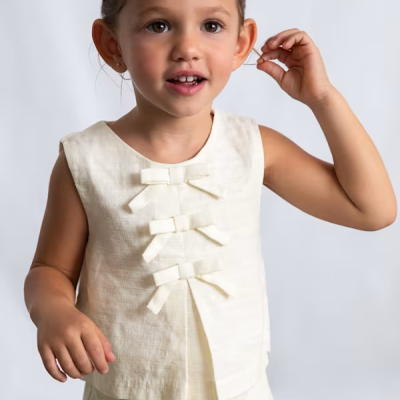 Conjunto infantil creme com blusa sem mangas e calças, detalhe de laços na blusa e cintura, em fundo claro.