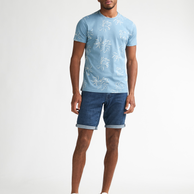 Homem com t-shirt azul com flores brancas, calções de ganga e sapatilhas brancas