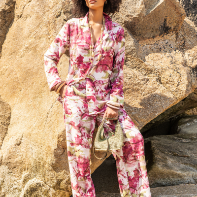 Mulher com conjunto floral rosa e branco, acessórios em madeira, bolsa de palha e óculos de sol em cenário rochoso