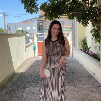 Mulher com vestido de riscas castanhas e brancas segurando mala branca em passagem exterior com plantas.