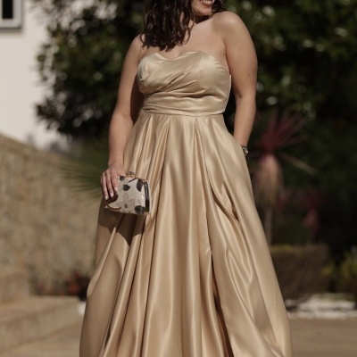Mulher de vestido dourado acetinado longo, com mala de mão floral e sandálias douradas em exterior ensolarado