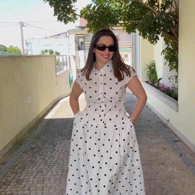 Vestido branco de bolinhas pretas com gola e botões, sandálias brancas, óculos escuros, mulher sorridente em passadeira de pedra.