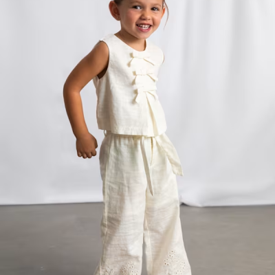 Menina sorridente com conjunto branco de top e calças largas com laços e bordados.