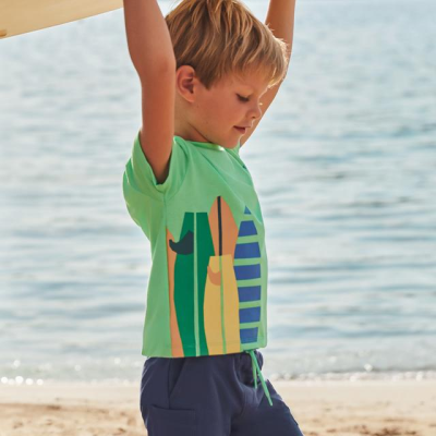Menino na praia com camisola verde clara e estampa de pranchas de surf, e calções azul escuro