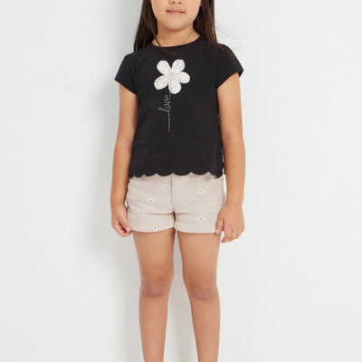 Menina com camisola preta de flor branca, calções bege com flores e sandálias douradas em fundo branco.