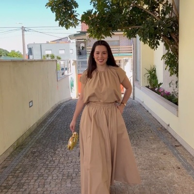 Mulher com conjunto bege e bolsa amarela em calçada de pedras sob árvore em ambiente urbano