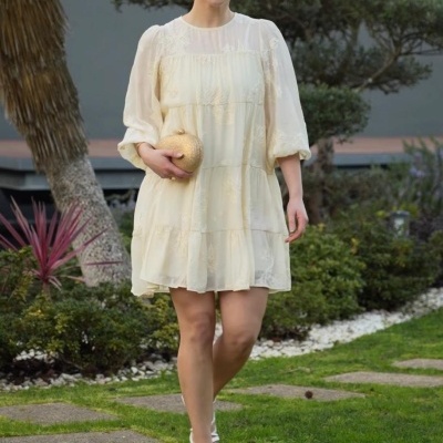 Mulher com vestido creme bordado, sandálias brancas e mala dourada num jardim