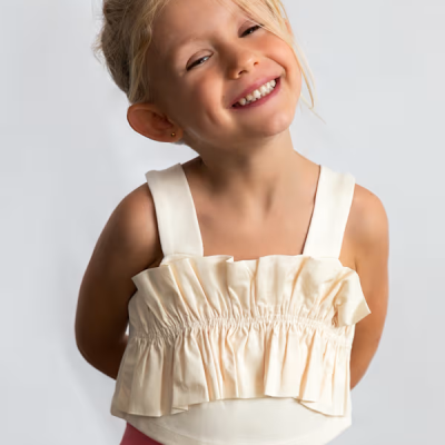 Menina loira sorridente com top creme franzido e calças cor-de-rosa com bolsos e botões