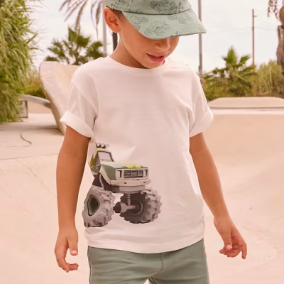 Menino com t-shirt branca estampada, calções verdes claros e boné verde no parque urbano com palmeiras