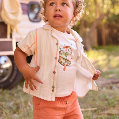 Menino com roupa clara e colorida ao ar livre com vegetação e caravana ao fundo