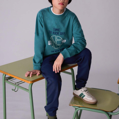Menino com camisola verde, calças azuis e sapatilhas brancas sentado em mesa e cadeira verde.