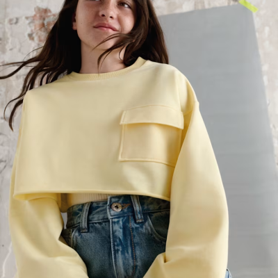 Sweatshirt cropped amarelo claro com bolso e calças de ganga azuis em ambiente com parede de tijolos