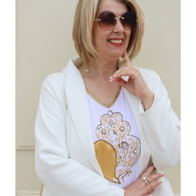 Mulher com óculos de sol, blazer branco e camisola com estampado dourado, fundo creme