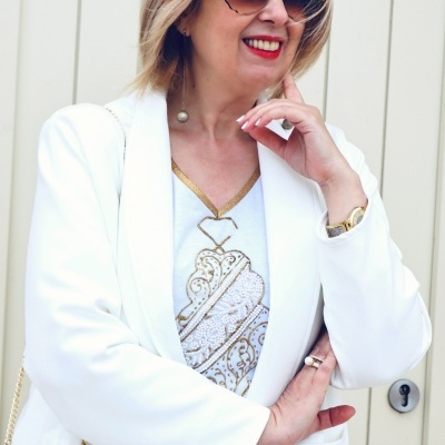 Mulher com óculos de sol flor, blazer branco e blusa branca com padrão dourado perto de porta clara