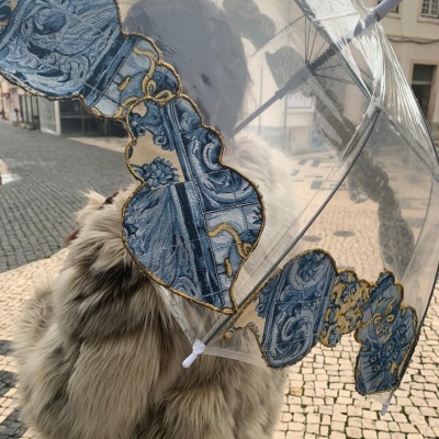 Guarda-chuva transparente com bordado azul e dourado, pessoa com casaco peludo bege, rua de calçada portuguesa