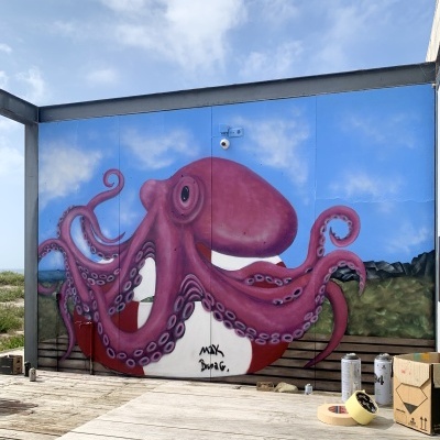 Mural de polvo roxo pintado em parede azul com nuvens, com assinatura MXK e Bruna G.