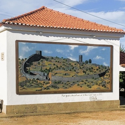 Mural pintado numa parede branca com aldeia antiga com muralhas e torres e céu azul