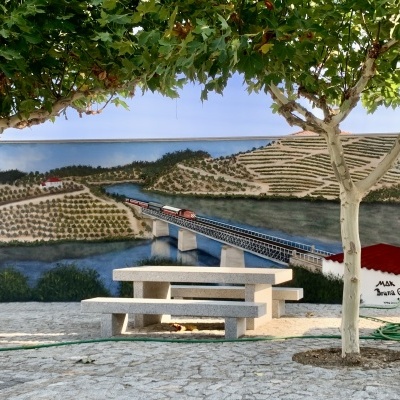 Banco de jardim de pedra com mesa ao ar livre e mural pintado de paisagem rural com ponte e comboio