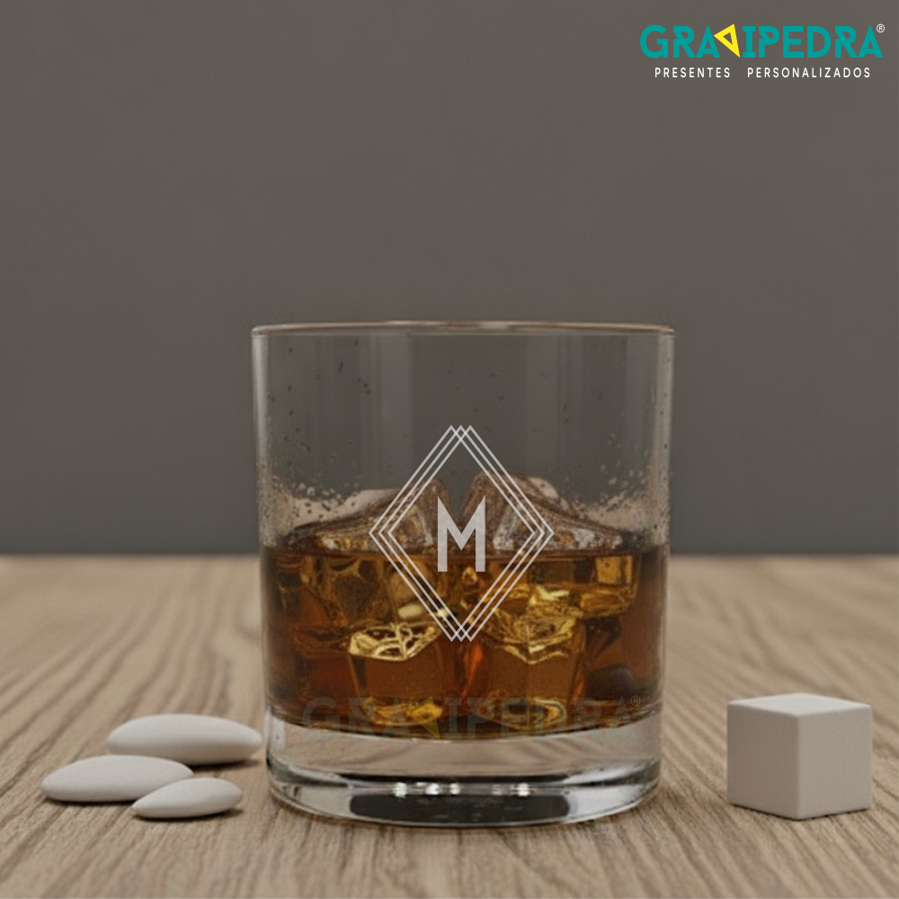 Copo em vidro personalizado - Inicial Nome - CVWD04 Copo de vidro com bebida castanha e gelo, decorado com letra M, sobre mesa de madeira clara com pedras brancas e cubo.