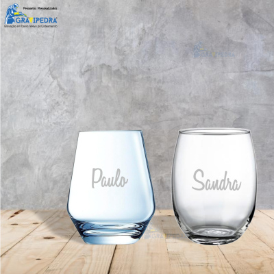 Dois copos de vidro com nomes 'Paulo' e 'Sandra' gravados, sobre superfície de madeira, fundo de parede de cimento