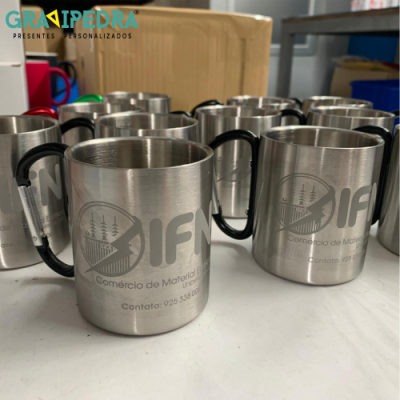 Canecas metálicas prateadas com logotipo CINAF e pega preta sobre mesa com caixas ao fundo.