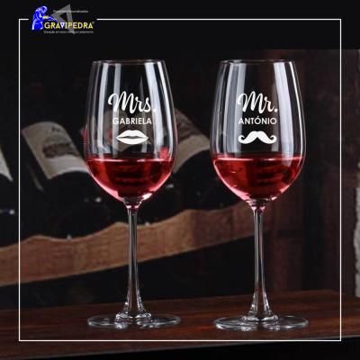Dois copos de vinho personalizados com nomes e desenhos, sobre mesa de madeira, fundo com garrafas de vinho