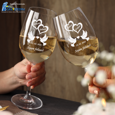 Dois copos de vinho branco gravados personalizados com nomes, data e mensagem, segurados em ambiente com fundo de madeira.