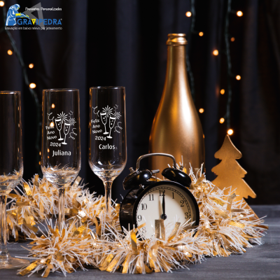 Copos de champanhe gravados com textos de ano novo, garrafa dourada, decoração dourada e relógio preto cercados por decoração festiva.