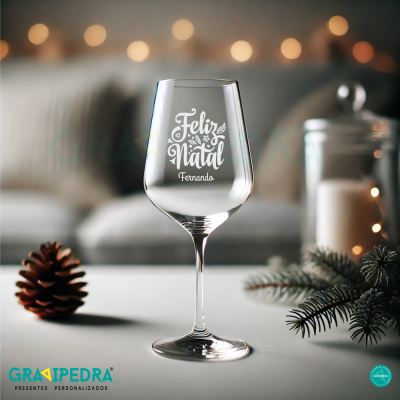 Taça de vinho com texto Feliz Natal Fernando, pinha e ramos de pinheiro numa mesa branca
