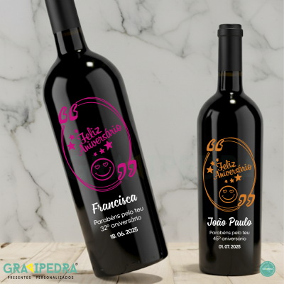 Duas garrafas de vinho tinto personalizadas para aniversários com textos em rosa e laranja, sobre mesa de madeira e fundo de mármore.