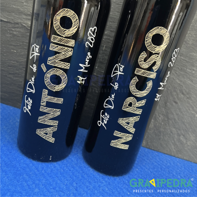 Duas garrafas pretas personalizadas com texto dourado e branco colocadas sobre superfície azul