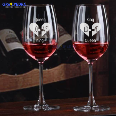 Dois copos de vinho com gravuras de corações e texto QUEEN KING LOVE KING QUEEN, vinho tinto dentro e adega ao fundo.