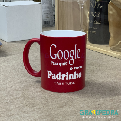 Caneca vermelha com texto branco 'Google Para quê? o meu Padrinho SABE TUDO' sobre mesa