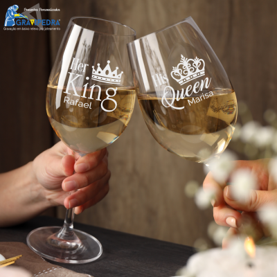 Duas taças de vinho branco gravadas com 'Her King Rafael' e 'His Queen Marisa', seguradas por duas mãos, com fundo desfocado