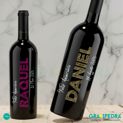 Garrafas de vinho tinto personalizadas com nomes RAQUEL e DANIEL e datas, sobre superfície branca marmoreada.